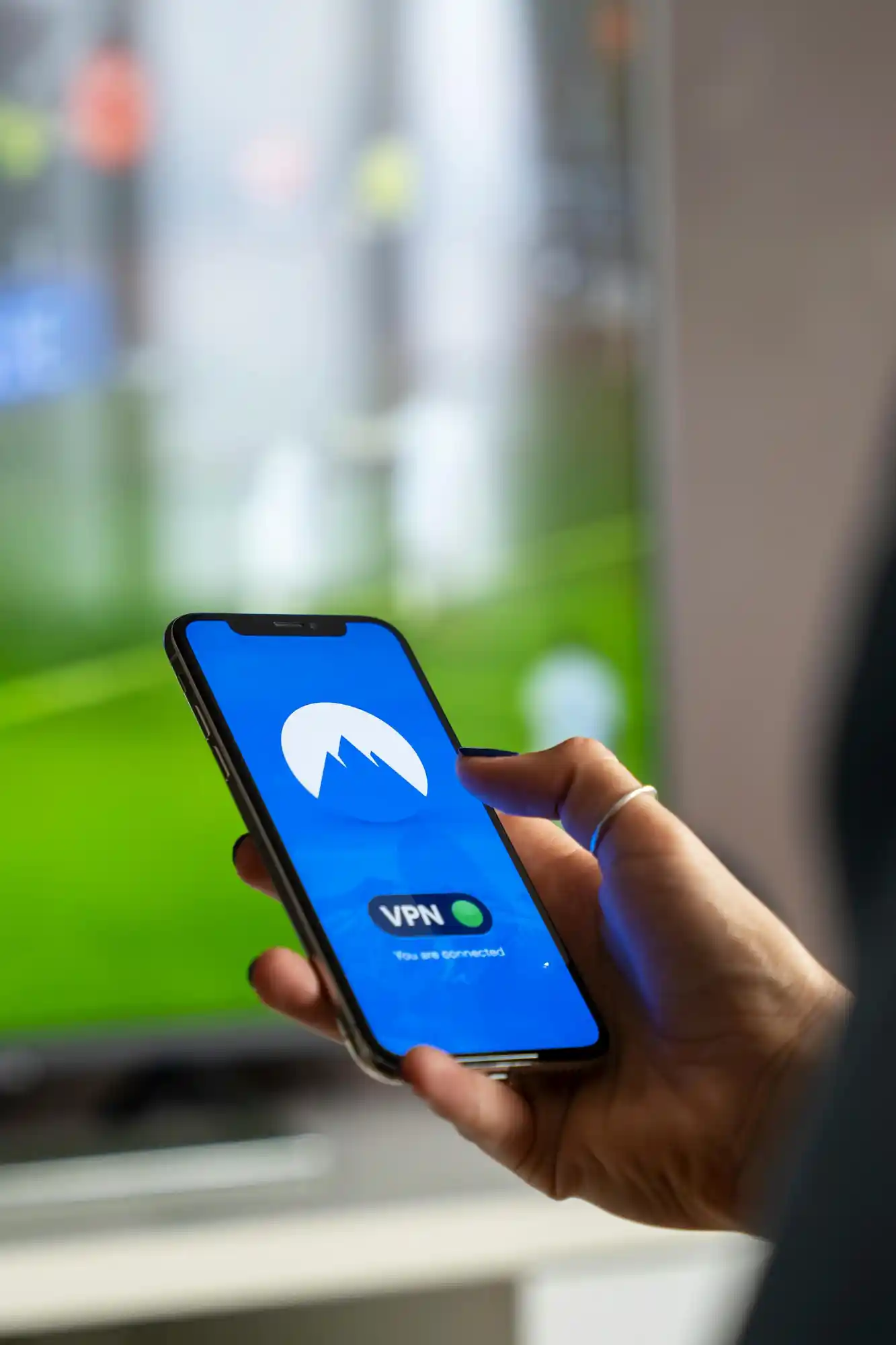 A VPN app logo is visible on the screen of a smartphone held in a hand. The background scene is somewhat blurred, suggesting that the user is turning on a privacy program or safe connection.